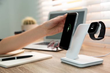 Bir kadın ahşap masada cep telefonunu kablosuz şarj cihazına bağlıyor. Modern işyeri aksesuarı