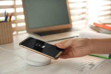 Beyaz ahşap masadaki cep telefonunu kablosuz şarj cihazına bağlayan kadın, yakın plan. Modern işyeri aksesuarı