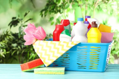 Bahar çiçekleriyle dolu plastik sepet ve açık mavi ahşap masada temizlik malzemeleri.