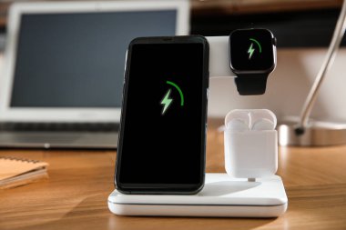 Cep telefonu, kulaklıklar ve ahşap masada kablosuz ped ile şarj olan akıllı sehpa, yakın plan. Modern işyeri aygıtı