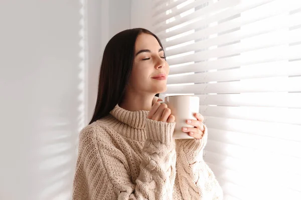 年轻女人穿着暖和的毛衣 在家里窗户边喝一杯热饮 — 图库照片