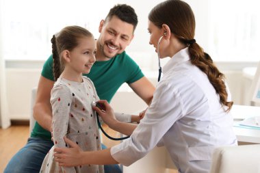 Baba ve kızı çocuk doktorunu ziyaret ediyor. Doktor, hastanede steteskoplu küçük bir hastayı muayene ediyor.