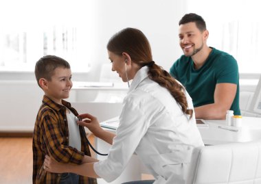 Baba ve oğul çocuk doktorunu ziyaret ediyor. Doktor, hastanede steteskoplu küçük bir hastayı muayene ediyor.