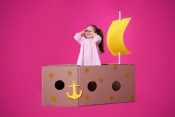 ピンクの背景に段ボール箱で作られた船で遊ぶ小さな子供 — ストック写真