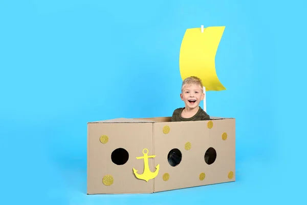 ライトブルーの背景に段ボール箱で作られた船で遊ぶ小さな子供 — ストック写真