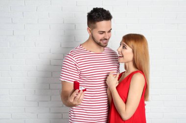 Nişan yüzüklü bir adam, beyaz tuğla duvarın yanında kız arkadaşına evlenme teklif ediyor.