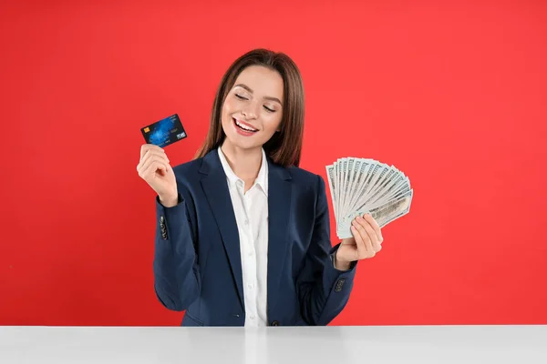 Young woman with money and credit card at table on crimson background