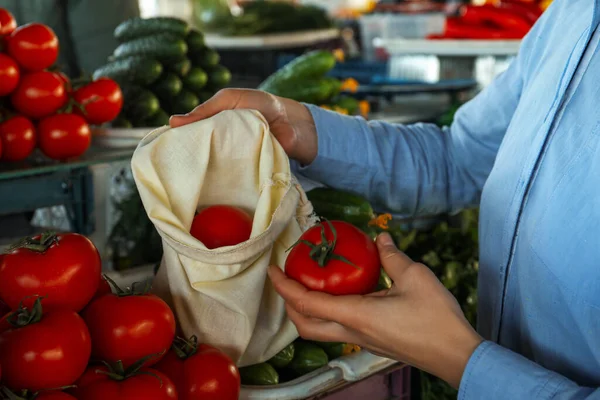 Woman putting tomato into cotton eco bag at wholesale market, closeup. Life without plastic