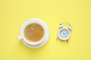 Sarı arka planda bir fincan sabah kahvesi ve çalar saat.
