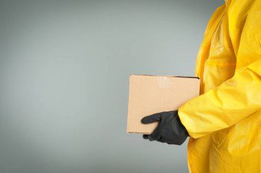 Kimyasal koruyucu giysili bir adam açık gri arka planda karton bir kutu tutuyor, yakın plan görüntüsü ve mesaj için yeri var. Virüsün yayılmasını önlemek