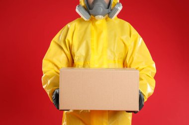 Kırmızı arka planda karton kutulu kimyasal koruyucu elbise giyen adam, yakın plan. Virüsün yayılmasını önlemek