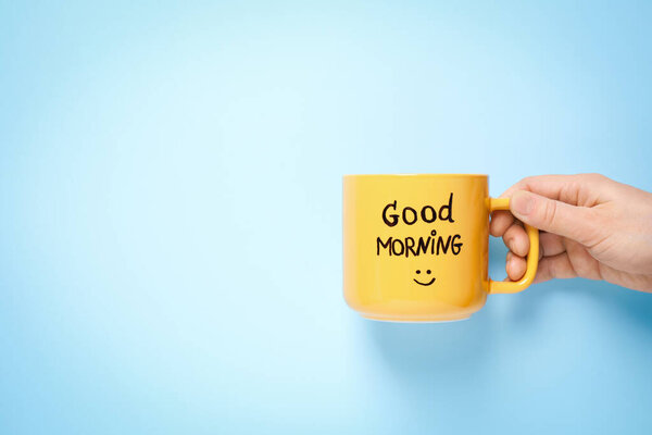 Woman holding cup of coffee with words GOOD MORNING on light blue background, closeup. Space for text
