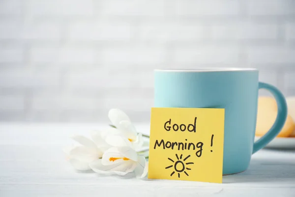 Delicious coffee, flowers and card with GOOD MORNING wish on white wooden table, closeup. Space for text
