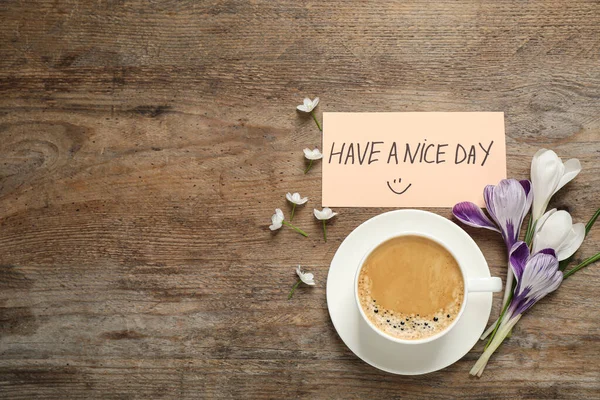 Morning coffee, flowers and card with HAVE A NICE DAY wish on wooden table, flat lay. Space for text
