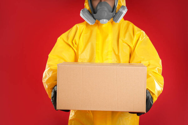 Man wearing chemical protective suit with cardboard box on red background, closeup. Prevention of virus spread