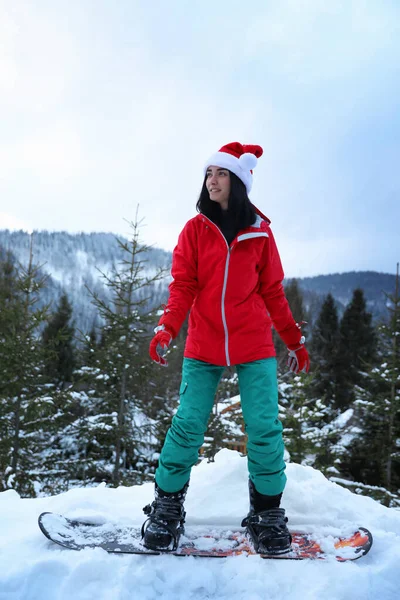 Kar tepesinde Noel Baba şapkası takan genç bir snowboardcu. Kış tatili