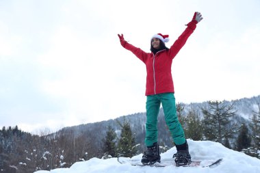 Kar tepesinde Noel Baba şapkası takan genç bir snowboardcu. Kış tatili
