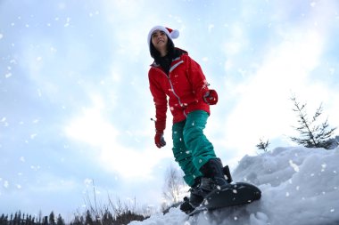 Kar tepesinde Noel Baba şapkası takan genç snowboardcu, alçak açılı. Kış tatili