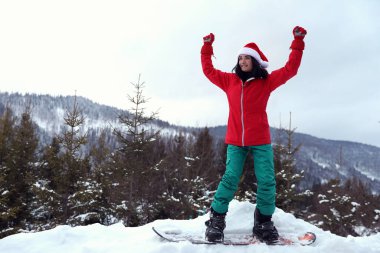 Kar tepesinde Noel Baba şapkası takan genç bir snowboardcu. Kış tatili