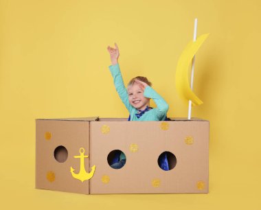 Küçük çocuk sarı arka planda karton kutudan yapılmış gemiyle oynuyor.