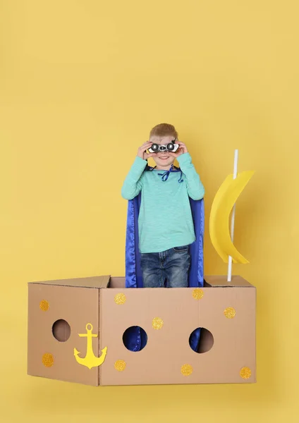 穿着斗篷的小孩玩黄色背景纸板箱做的船 — 图库照片