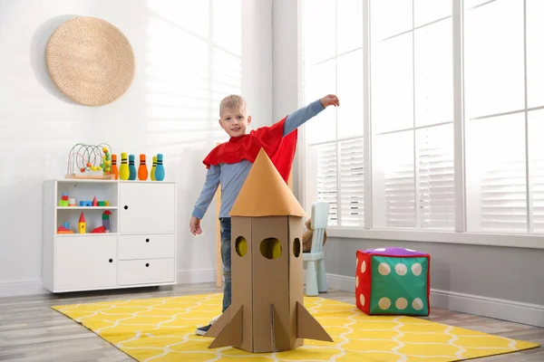 家で段ボール箱で作られたロケットで遊ぶ赤いケープの小さな子供 — ストック写真