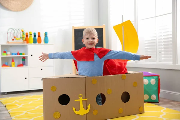 家で段ボール箱で作られた船と遊んで赤いケープの小さな子供 — ストック写真
