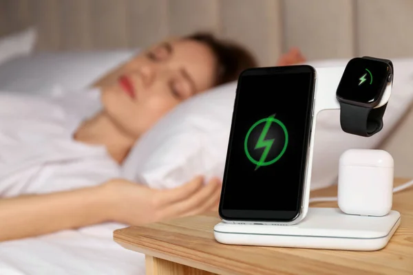 Smartphone, watch, earphones charging on wireless pad and sleeping woman in room