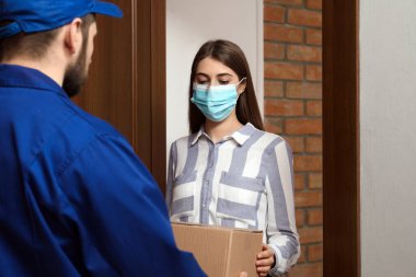 Tıbbi maske takan genç bir kadın içeride teslimatçıdan paket alıyor. Virüsün yayılmasını önlemek