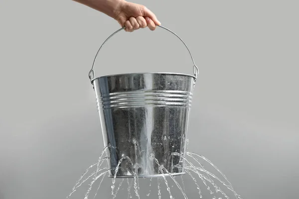 Woman holding leaky bucket with water on light grey background, closeup