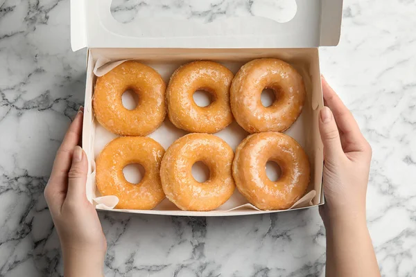 Woman with box of delicious donuts at marble table, top view