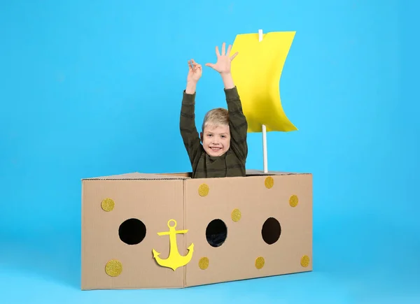 ライトブルーの背景に段ボール箱で作られた船で遊ぶ小さな子供 — ストック写真
