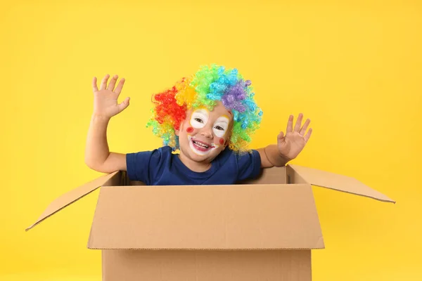 戴小丑假发的小男孩坐在黄色背景的纸板箱里 四月愚人节 — 图库照片