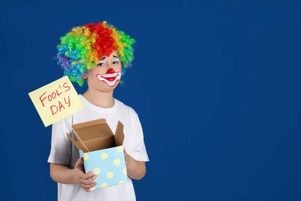 身穿小丑装束头戴假发的十多岁男孩手持蓝色背景的 四月愚人节 文字空间 — 图库照片