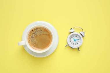 Sarı arka planda bir fincan sabah kahvesi ve çalar saat.