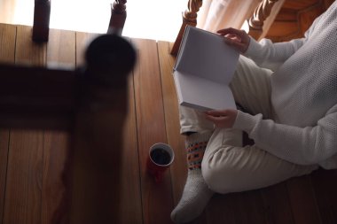 Elinde kahve fincanı olan bir kadın evde kitap okuyor.