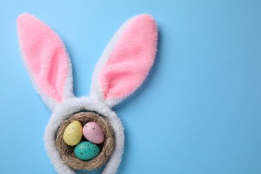 Paskalya tavşanı kulaklı saç bandı ve açık mavi arka planda yuvada boyanmış yumurtalar, düz yatak. Metin için boşluk
