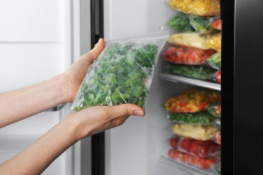 Buzdolabının yanında donmuş yeşil fasulyeli plastik poşet taşıyan bir kadın var.