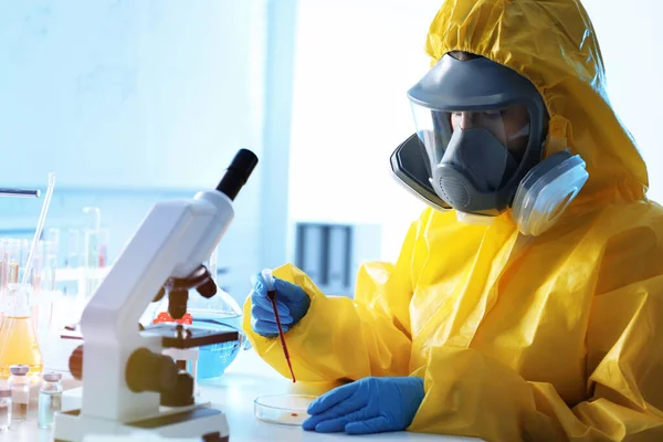 Scientist in chemical protective suit dripping blood sample into Petri dish at laboratory. Virus research