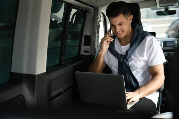 Handsome man with laptop talking on phone in modern car