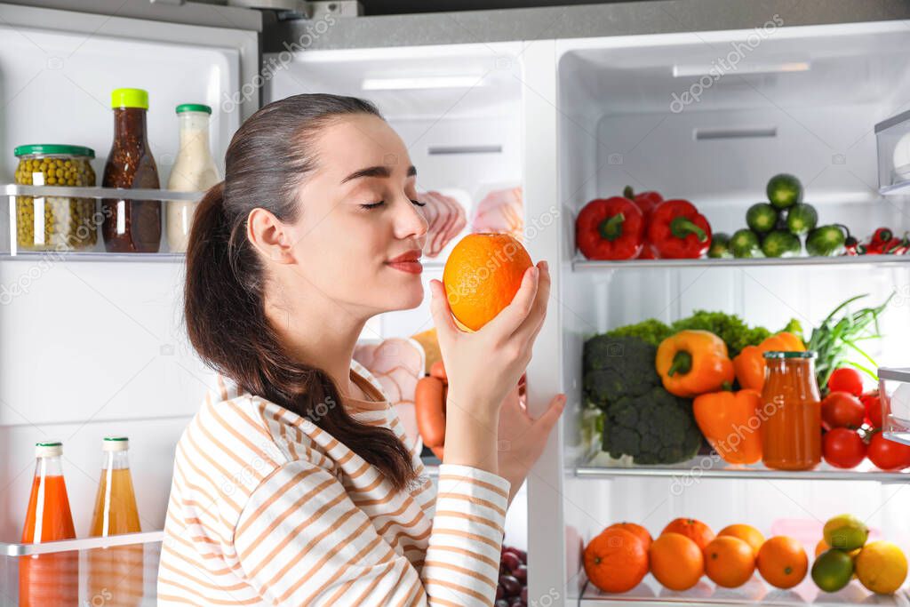 Young woman with orange near open refrigerator indoors