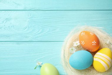 Dekoratif yuvada renkli Paskalya yumurtaları ve açık mavi ahşap arka planda çiçekler. Metin için boşluk