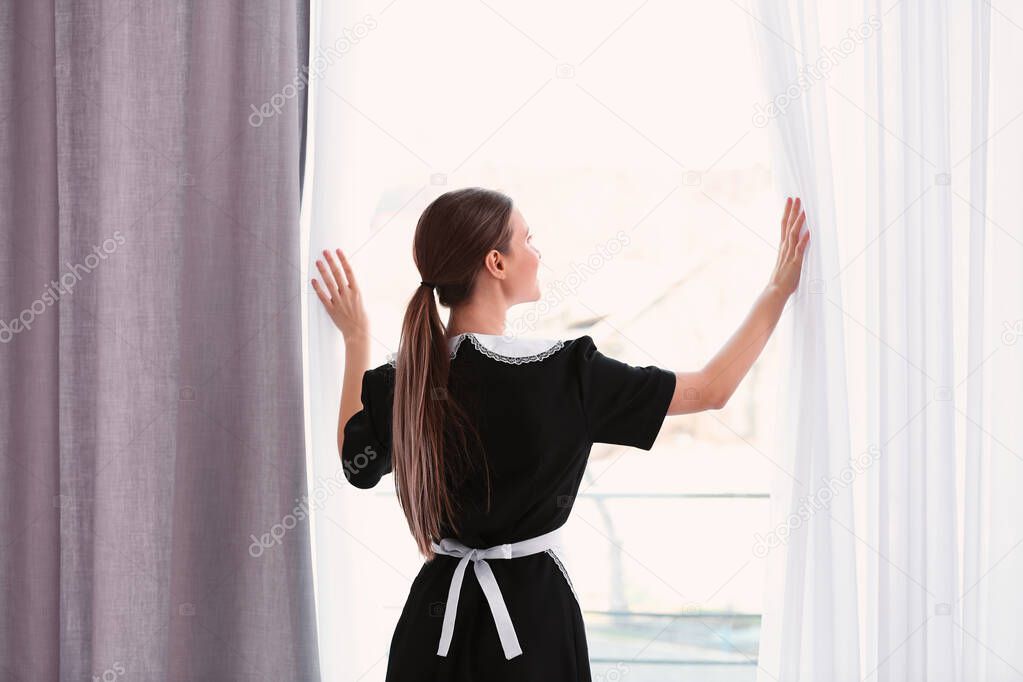 Young chambermaid opening window curtains in hotel room