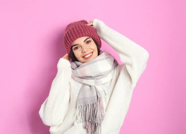 Pembe arka planda kalın kazak, eşarp ve şapka giyen genç bir kadın. Kış mevsimi