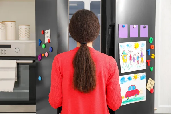 Woman opening refrigerator door with child\'s drawings, notes and magnets in kitchen