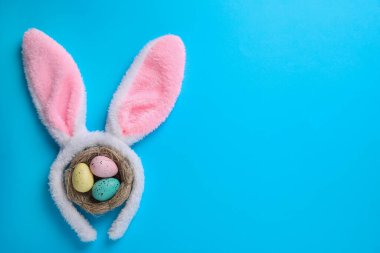 Paskalya tavşanı kulaklı saç bandı ve açık mavi arka planda yuvada boyanmış yumurtalar, düz yatak. Metin için boşluk