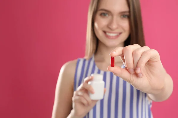 Young woman with vitamin pill against pink background, focus on hand