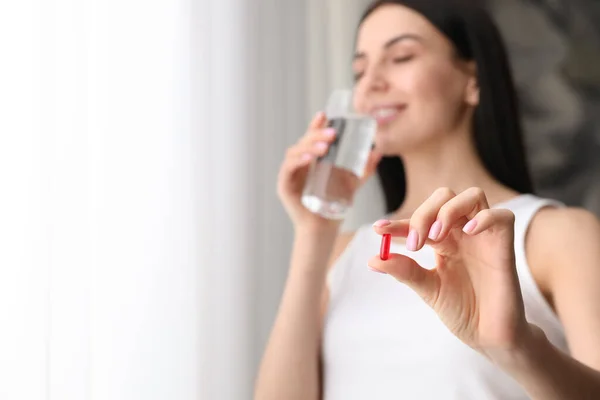 Woman with vitamin capsule and glass of water indoors, focus on hand. Space for text