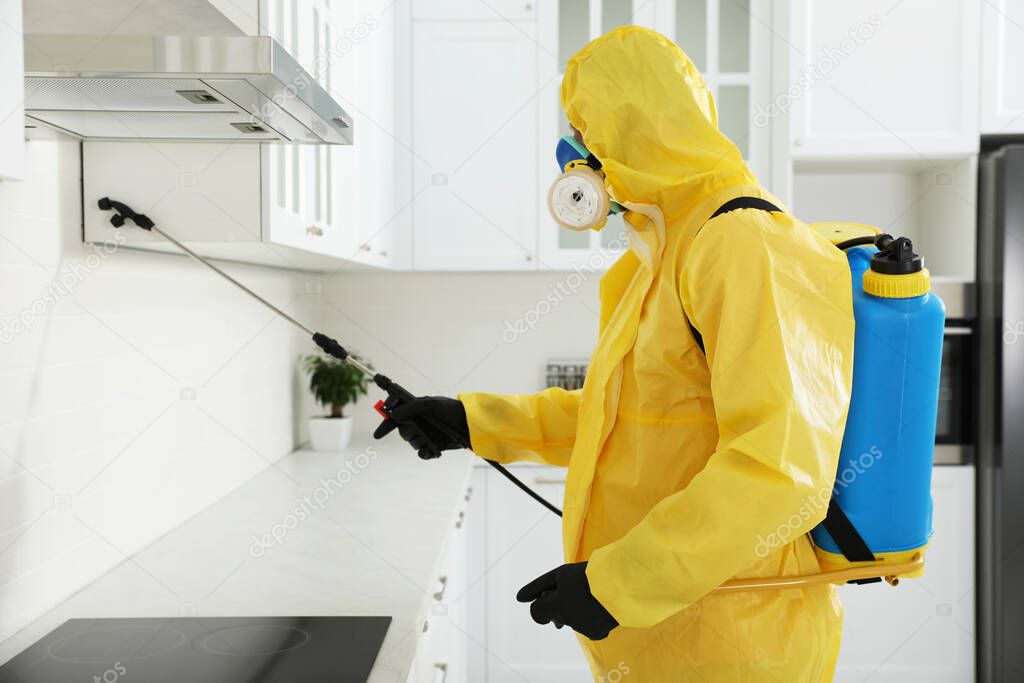 Pest control worker in protective suit spraying insecticide on furniture indoors