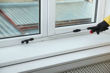 Koruyucu kıyafetli haşerat kontrol işçisi pencere pervazına böcek ilacı sıkıyor, yakın plan.